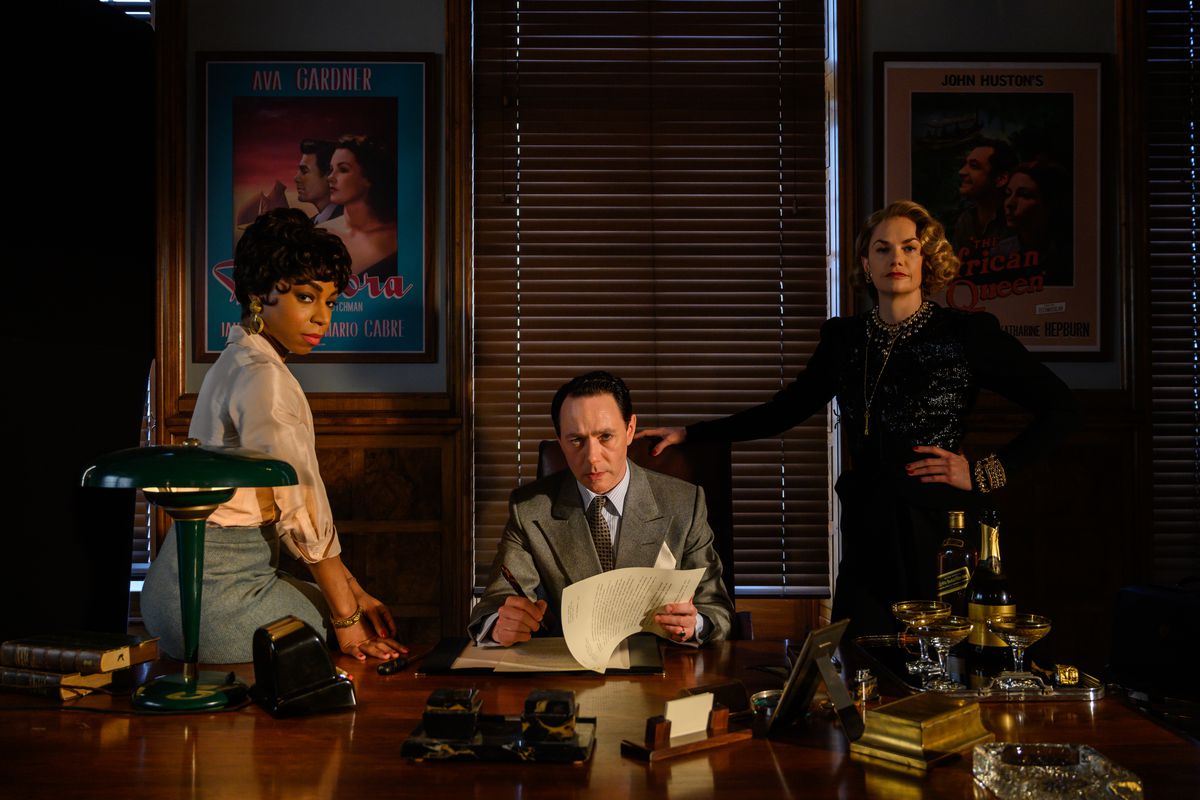 John Wolfe (Rice Shersmith), Petola Spencer (Rita Wilson) i Anne Savile (Peppa Bennett Warner) wychodzą zza dużego, błyszczącego biurka Wolfe'a.