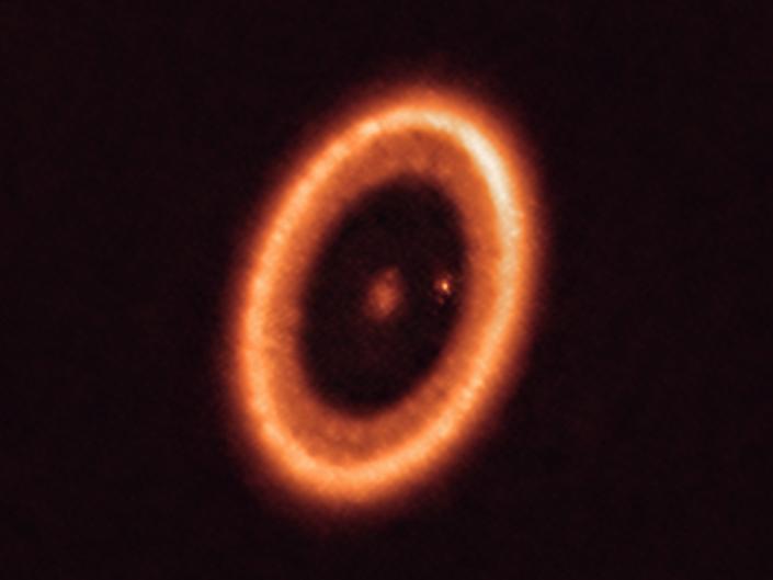 Obraz przedstawia pomarańczową gwiazdę otoczoną pomarańczowym dyskiem materii z małą planetą w kropki